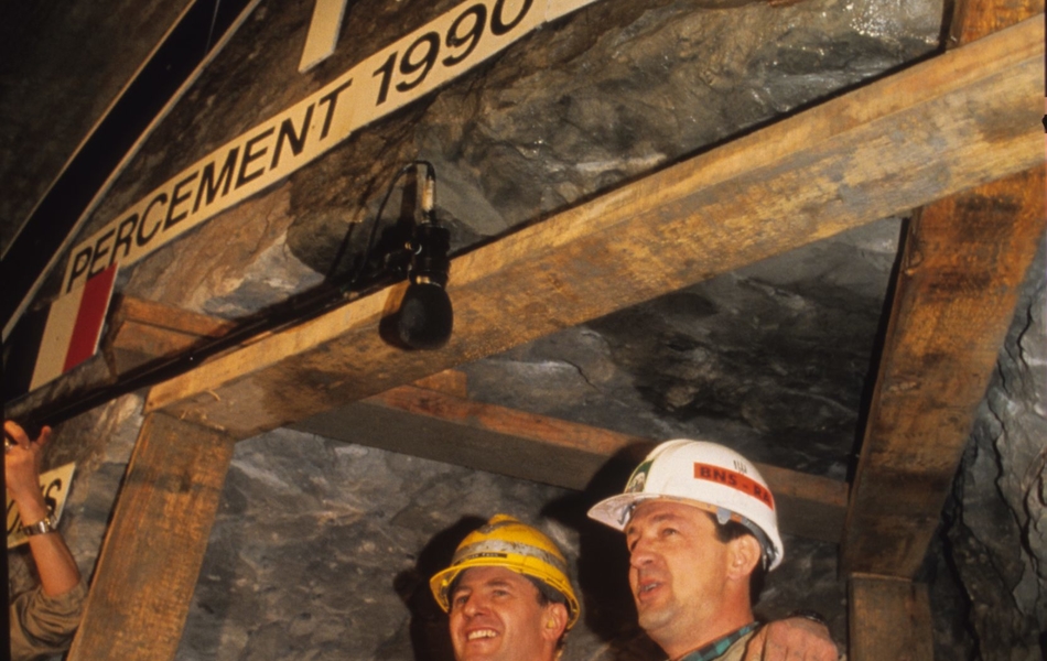 Photographie couleur montrant deux hommes souriant, en tenue d'ouvrier, se donnant une accolade.