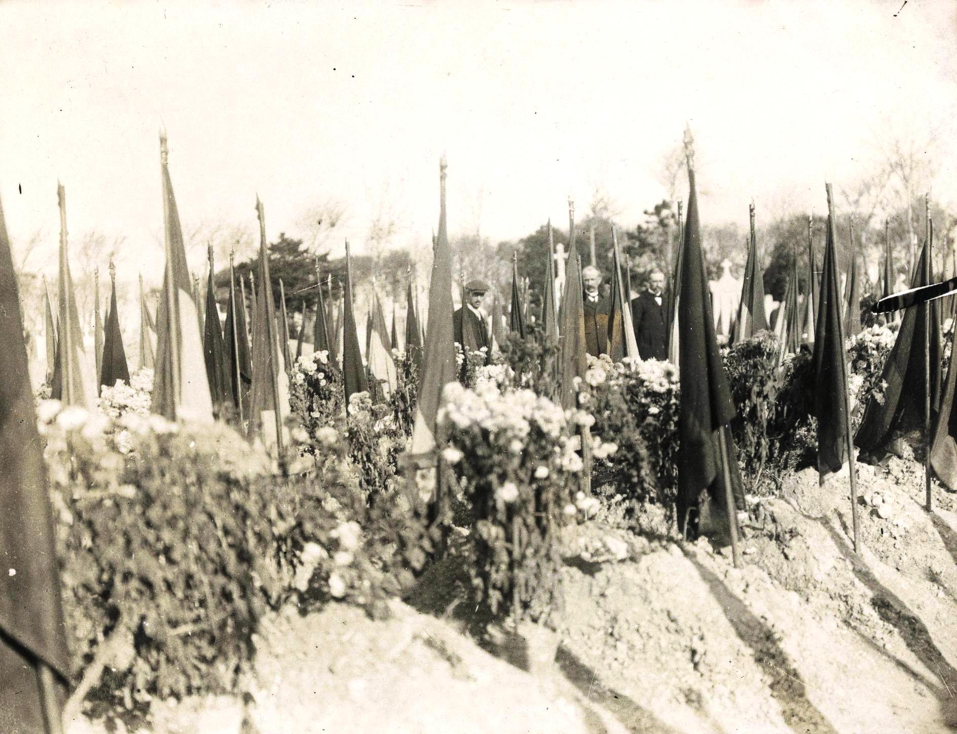 Photographie noir et blanc montrant des drapeau en berne dans un cimetière dans lequel se recueillent des personnes.