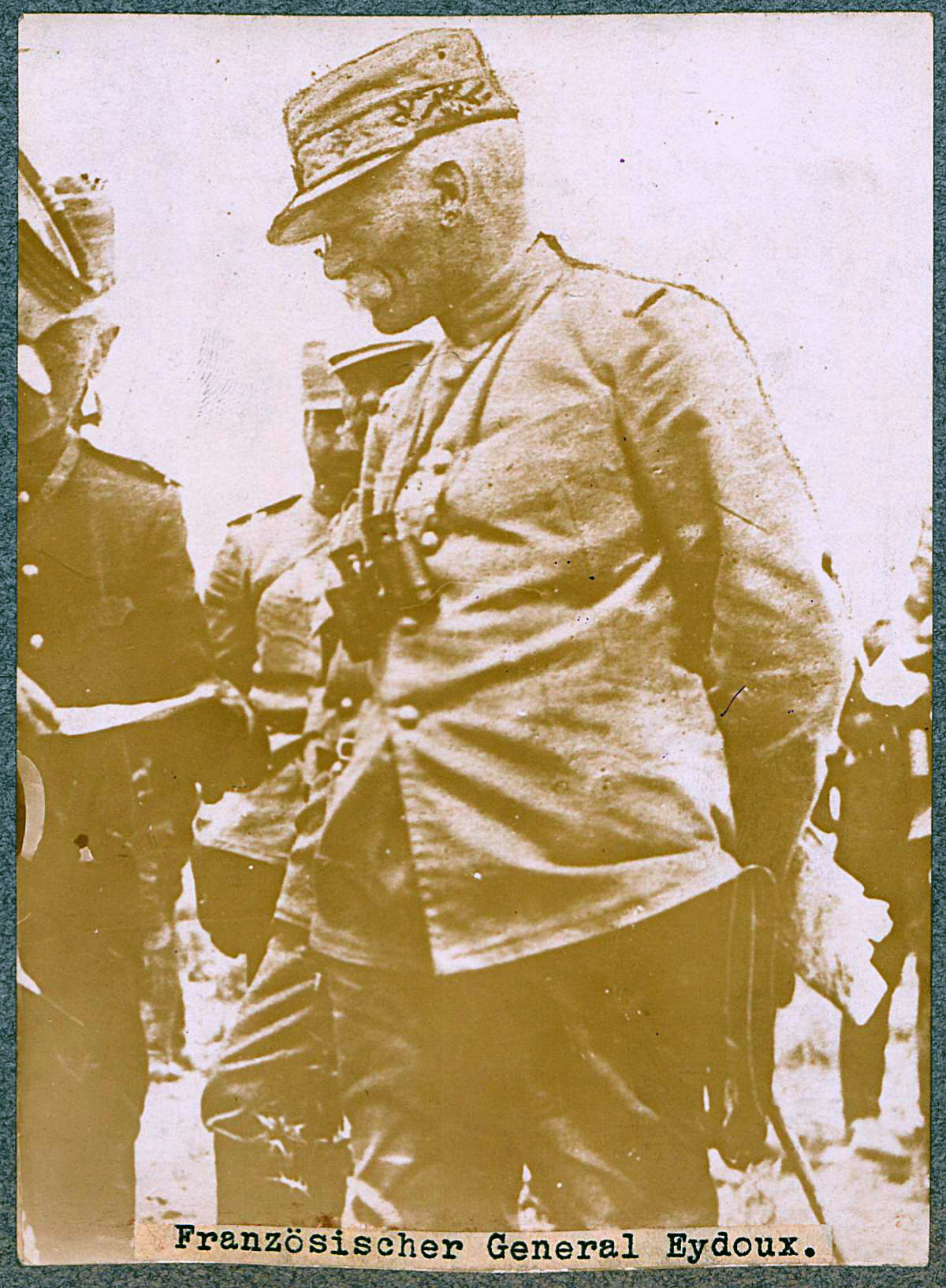 Photographie sepia montrant un homme de profil, en uniforme militaire, les mains dans le dos.