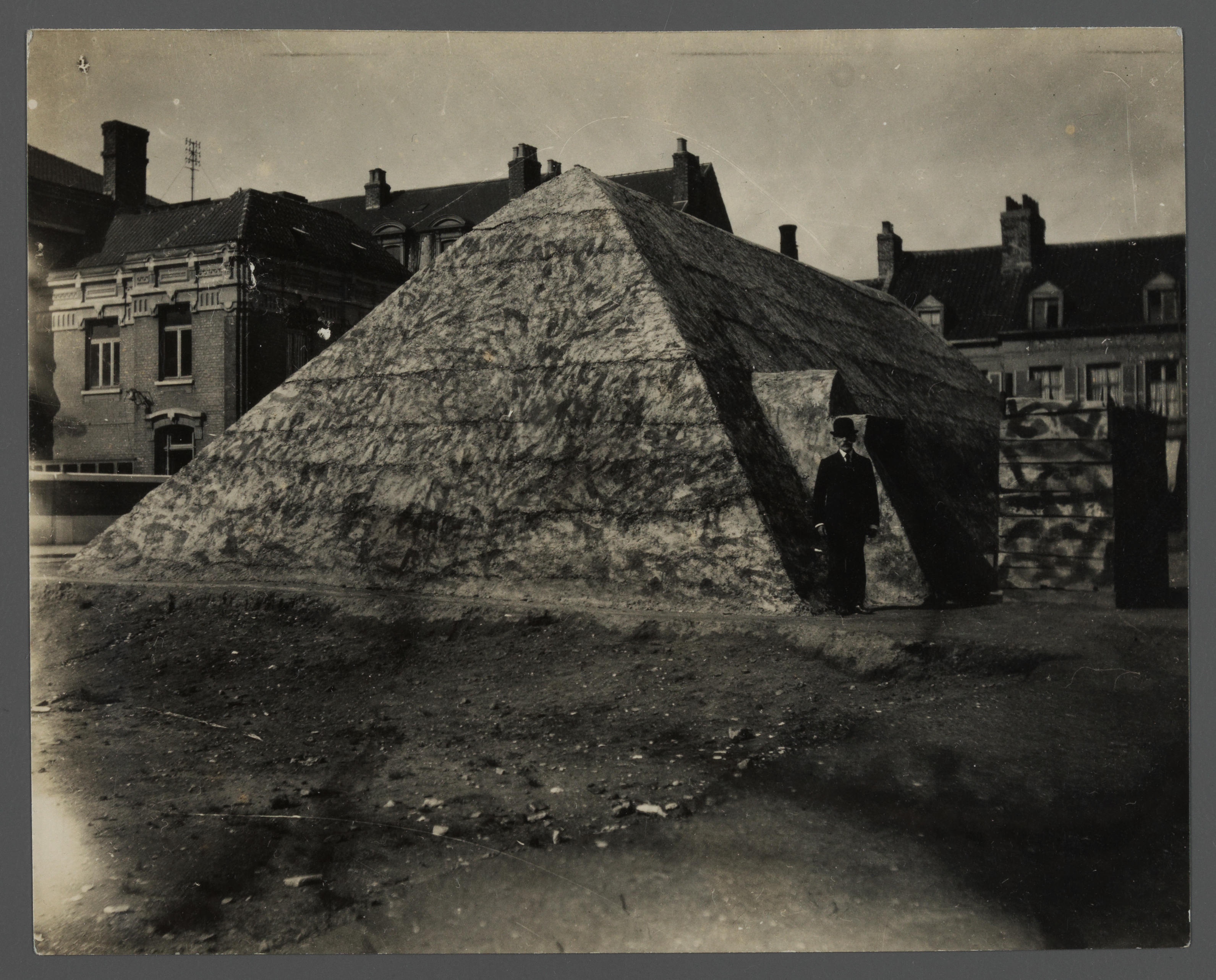 Photographie noir et blanc d'un abri en forme de prisme triangulaire sur une place. Un homme en costume pose à l'entrée.