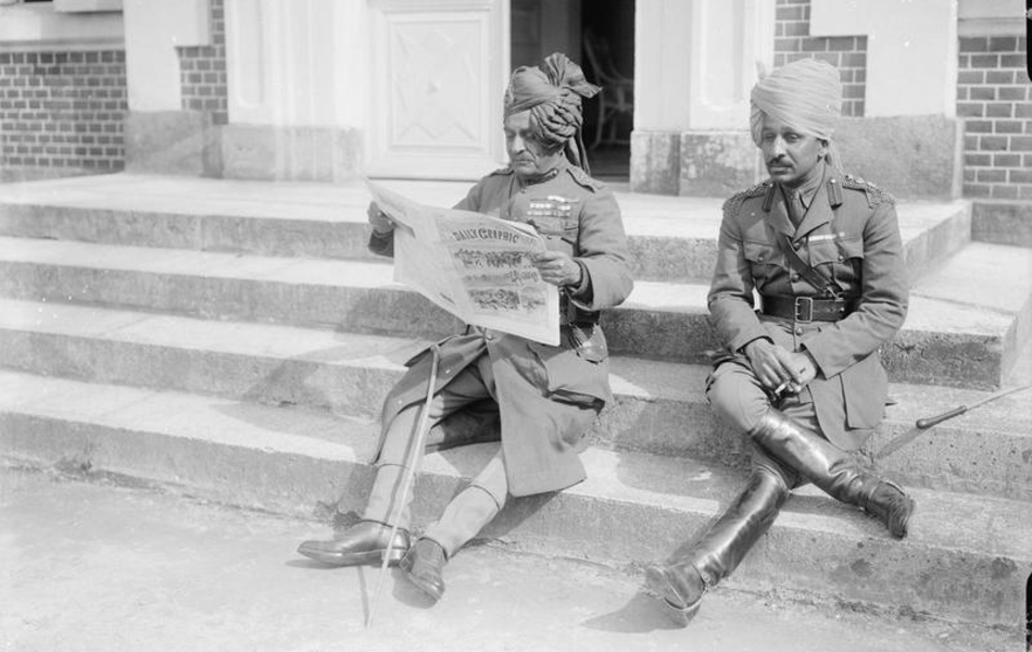 Photographie noir et blanc montrant deux soldats indiens assis sur les marches d'un perron. L'un fume tandis que l'autre lit le journal.