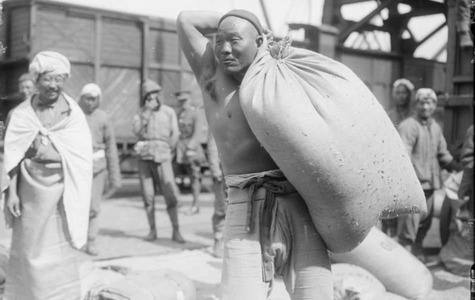 Photographie noir et blanc montrant au premier plan un asiatique, torse nu, portant un sac sur son épaule. À l'arrière plan, d'autres hommes devant un wagon de marchandises.