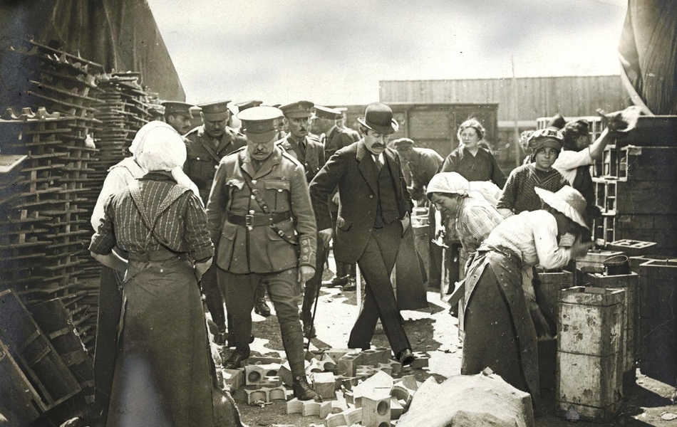 Photograhie noir et blanc montrant un groupe de militaires traversant un chantier sur lequel travaillent des femmes.