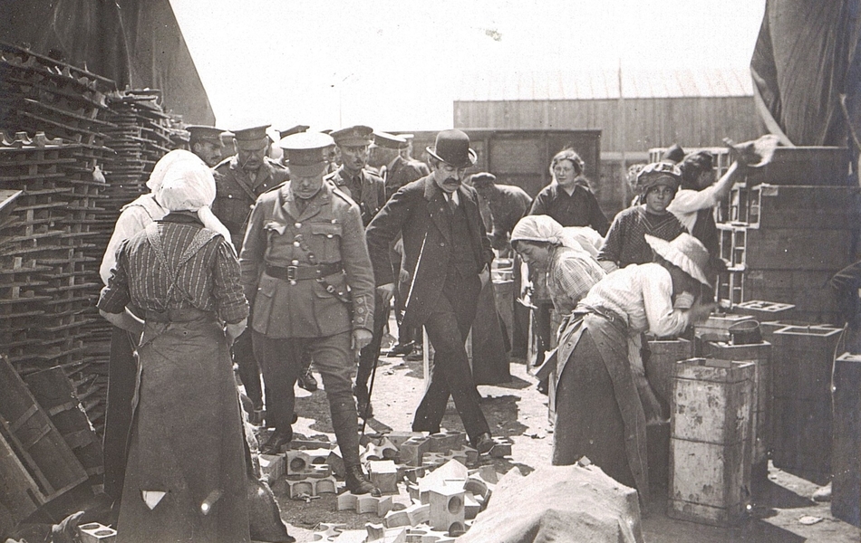 Photograhie noir et blanc montrant un groupe de militaires traversant un chantier sur lequel travaillent des femmes.