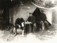 Photgraphie noir et blanc montrant trois hommes installés devant une tente. L'un semble lire une lettre pendant qu'un second rédige un courrier.