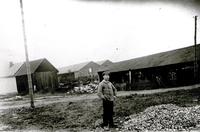 Photographie noir et blanc montrant un asiatique posant devant des hangars à la campagne.