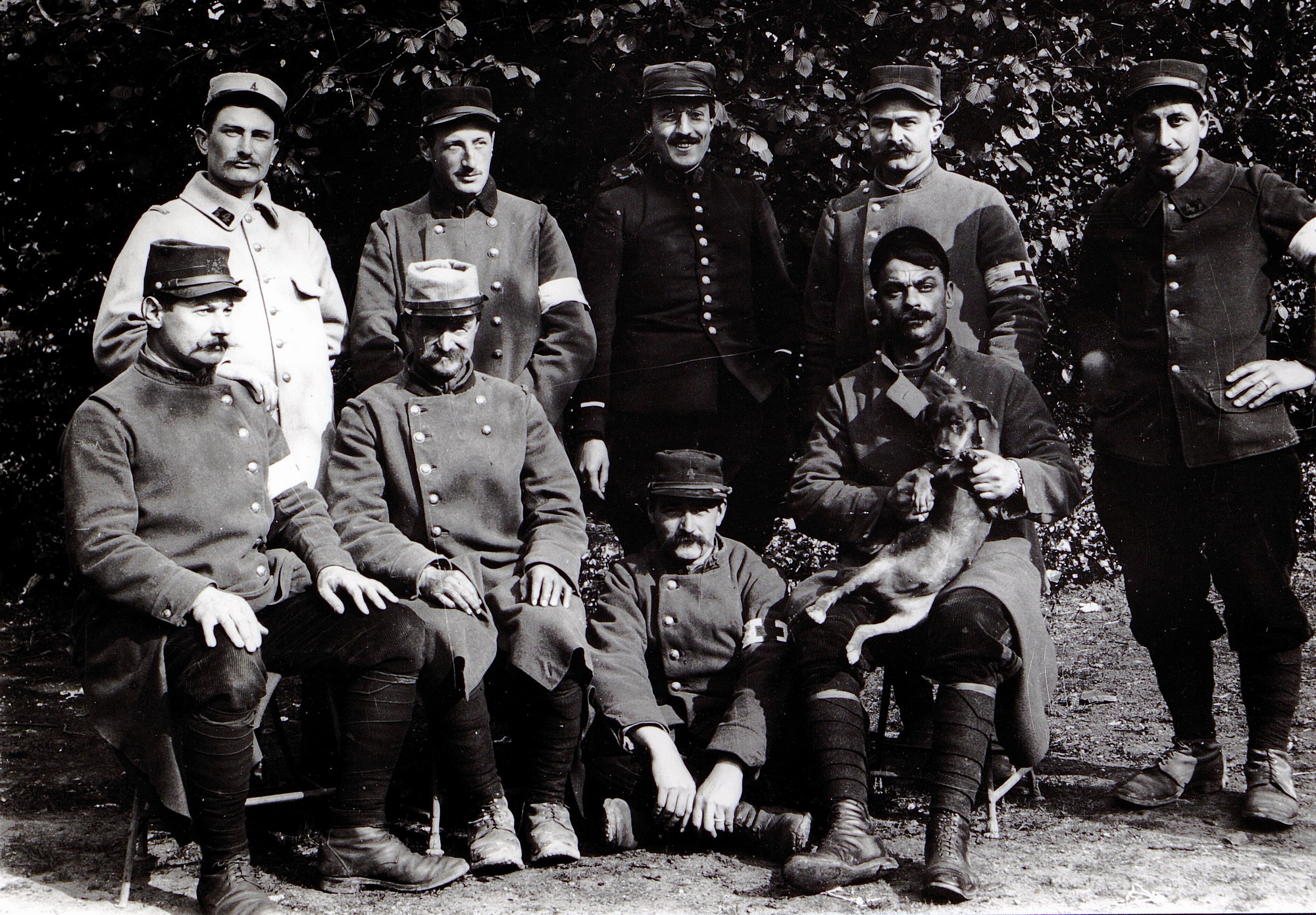 Photographie noir et blanc montrant un groupe de 9 soldats posant pour le photographe. L'un d'entre eux tient un chien sur ses genoux.