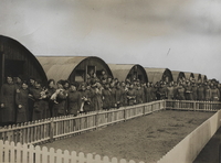 Photographie noir et blanc montrant un groupe de femmes posant devant des baraquements en tôle. 