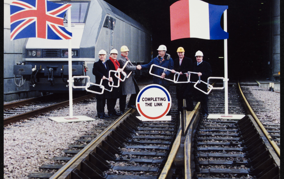Photographie couleur montrant des hommes portant des casques de chantier, réunis sur une voie ferrée à l'entrée d'un tunnel.