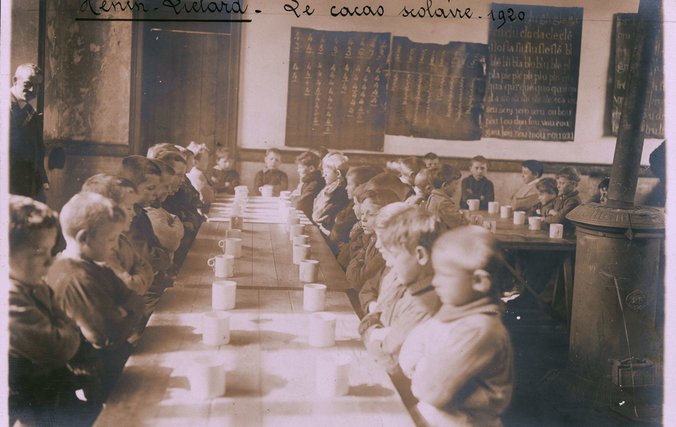 Photographie noir et blanc montrant des élèves dans un réfectoire, attablés devant une tasse de lait.