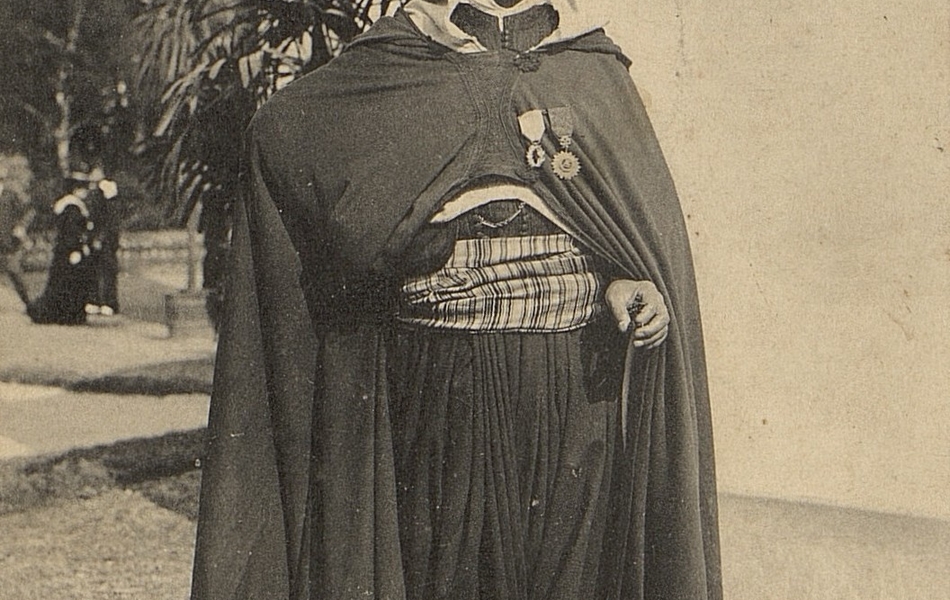 Carte postale noir et blanc d'un homme posant en pied, vêtu d'un costume traditionnel algérien et tenant un sabre. Sur le revers de sa cape sont épinglées deux médailles.