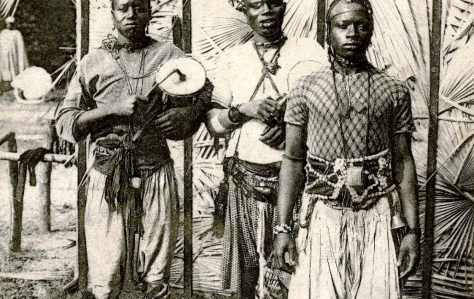 Carte postale noir et blanc montrant trois africains vêtus de costumes traditionnels. L'un d'entre eux porte une petite percussion.
