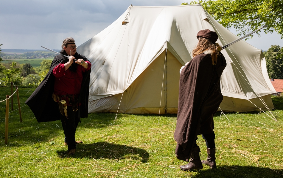 Photographie couleur montrant deux hommes vêtus à la mode du Moyen Age en train de se battre à l'épée devant une tente de campement.