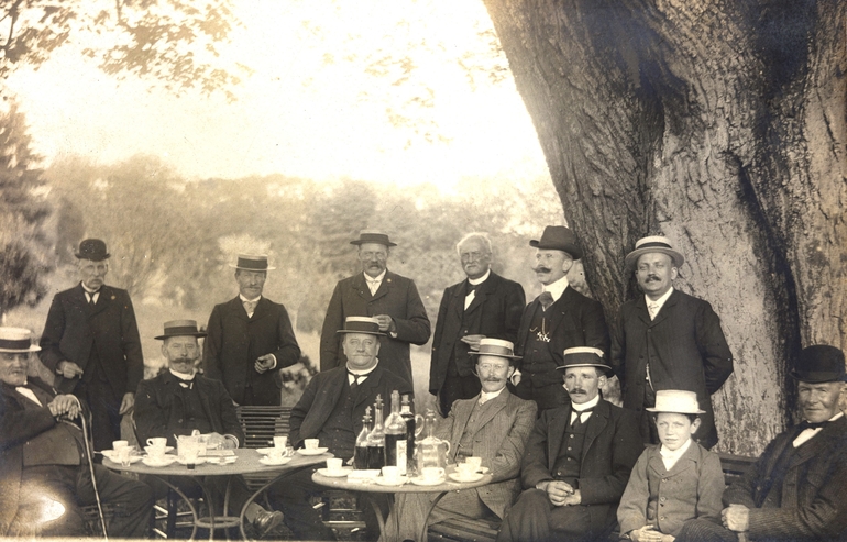 Photographie noir et blanc montrant un groupe de personnes déjeunant sous un arbre.