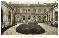 Carte postale noir et blanc montrant un cour intérieure avec parterre. Une fenêtre ouverte dans le bâtiment du fond laisse apercevoir cinq silhouettes féminines.