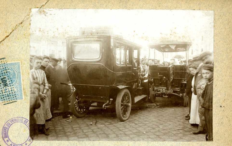 Photographie noir et blanc montrant, de derrière, deux voitures accidentées. Autour s'est rassemblée une grande foule ; les personnes se trouvant au premier plan regardent en direction du photographe. À gauche de la photographie, se trouve un timbre fiscal.