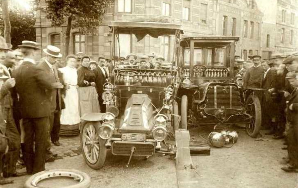 Photographie noir et blanc montrant, de devant, deux voitures accidentées. Autour s'est rassemblée une grande foule ; les personnes se trouvant au premier plan regardent en direction du photographe. 