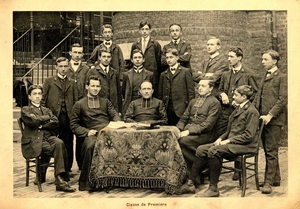 Photographie de treize élèves de première, en costume, dans la cour de l’institution Saint-Joseph, à Arras. Ils sont accompagnés par trois de leurs professeurs, en soutane et rabat, assis au premier rang devant une petite table nappée, où sont posés trois livres.