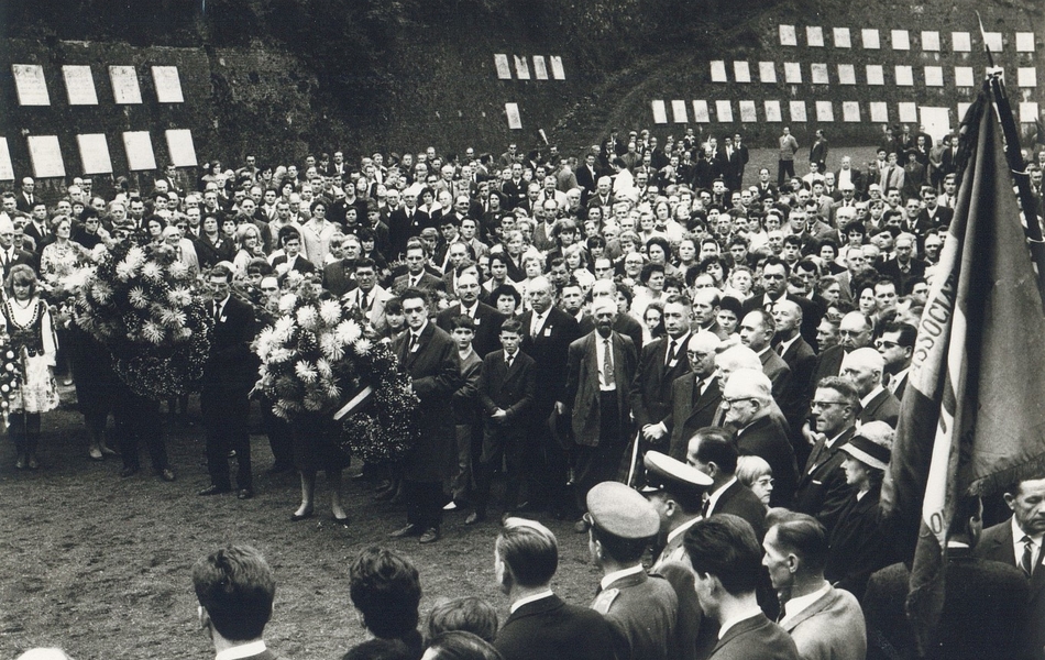 Photographie noir et blanc montrant une foule rassemblée devant un mur portant des plaques. Certaines personnes portent des bouquets de fleurs.