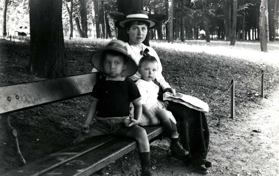 Photographie noir et blanc montrant une femme et deux jeunes enfants assis sur un banc dans un parc.