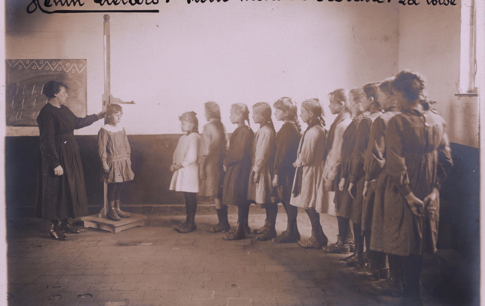 Photographie noir et blanc montrant des jeunes filles en file indienne qui sont mesurées à tour de rôle par une institutrice.