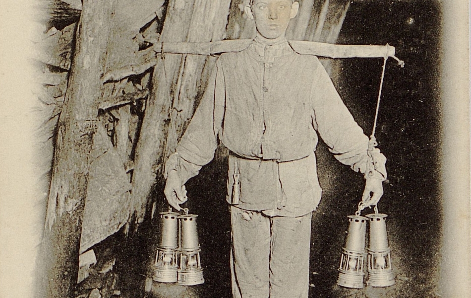 Carte postale noir et blanc montrant un jeune garçon portant des lampes de mineur dans une galerie de mine.
