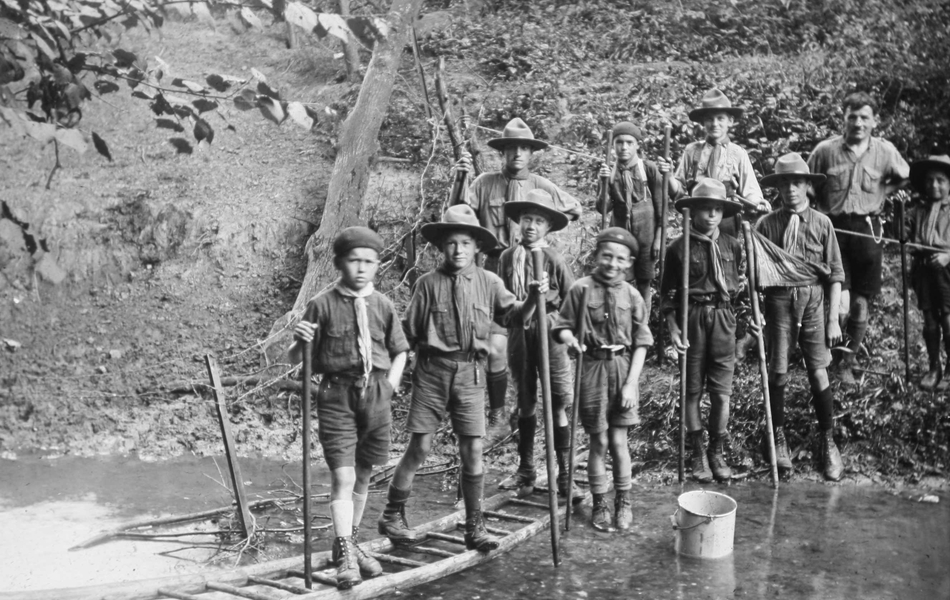 Photographie noir et blanc montrant de jeunes scouts en train de franchir un torrent grâce à un pont improvisé avec une échelle.