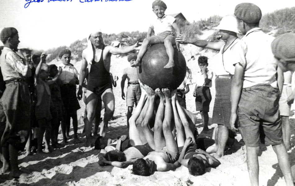 Photographie noir et blanc montrant des garçonnets sur une plage. Quelques uns d'entre eux soutiennent un petit garçon assis sur un ballon en caoutchou.