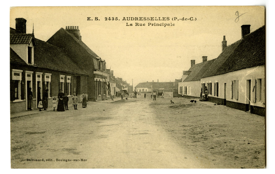 Carte postale en noir et blanc d’une large rue non pavée, bordée de petites maisons mitoyennes. Plusieurs personnes, debout devant leurs portes, regardent en direction du photographe