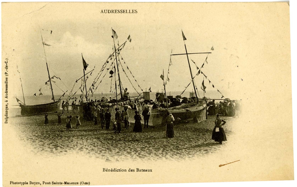 Carte postale en noir et blanc intitulée "Bénédiction des bateaux" représentant plusieurs barques de pêcheurs sur la plage encadrées d’une foule. Leurs mâts sont décorés de fanions et, derrière, on distingue des oriflammes