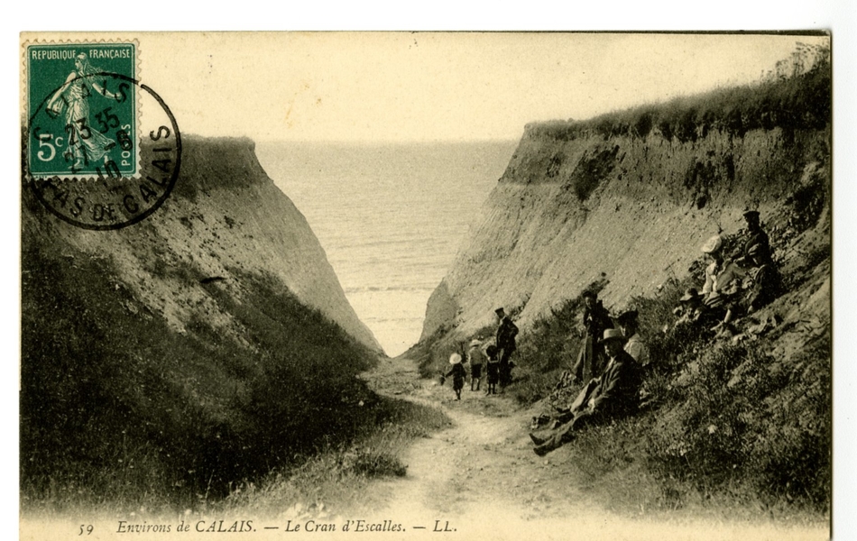 Carte postale en noir et blanc d’un sentier descendant vers la plage et entouré de falaises. Trois enfants accompagnés d’un homme se dirigent vers la mer, tandis qu’un groupe se repose sur le bas-côté