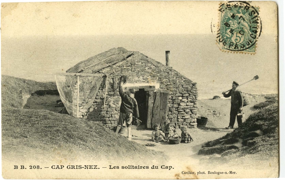 Carte postale en noir et blanc d’une cabane de pêcheur en pierres, au bord de la mer. Quatre enfants sont assis devant, autour d’un panier. Un homme pieds nus, le pantalon relevé et portant un grand chapeau de pêcheur, accroche des cordages au mur de la cabane, à côté d’une grande épuisette ; un deuxième homme, portant seau et panier en osier en bandoulière, rejoint le groupe