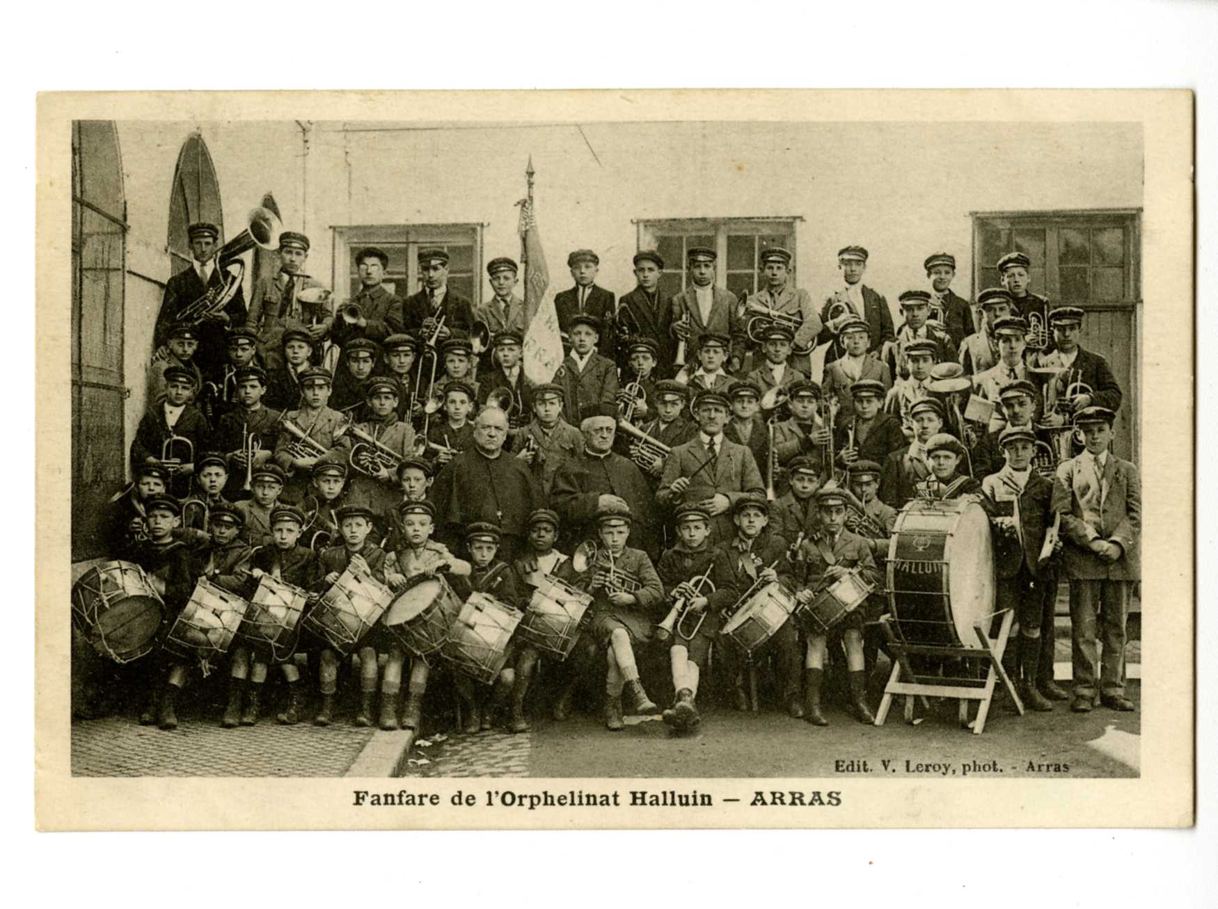Photographie noir et blanc de l'ensemble de la fanfare composée d'une trentaine d'enfants de tous âges, vêtus de leur uniforme et tenant leurs instruments. Au centre, trois hommes et derrière eux, la bannière.