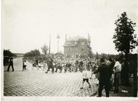 Photographie noir et blanc de soldats défilant dans une rue pavée sous le regard des badauds. En tête de cortège, on remarque des tambours puis des cuivres.