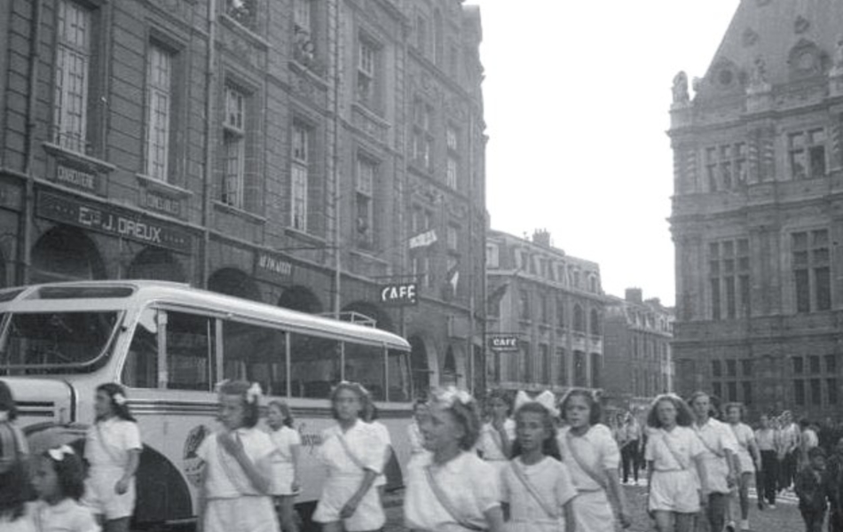 Photographie noir et blanc montrant un défilé de jeunes filles vêtues de blanc sur une place pavée.