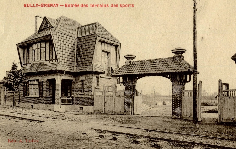 Carte postale noir et blanc d'une maison en briques prolongée latéralement par un porche ouvert ; à l'arrière plan, on distingue un terrain de sport, au premier plan les rails d'un train ou d'un tramway sur une route de terre.