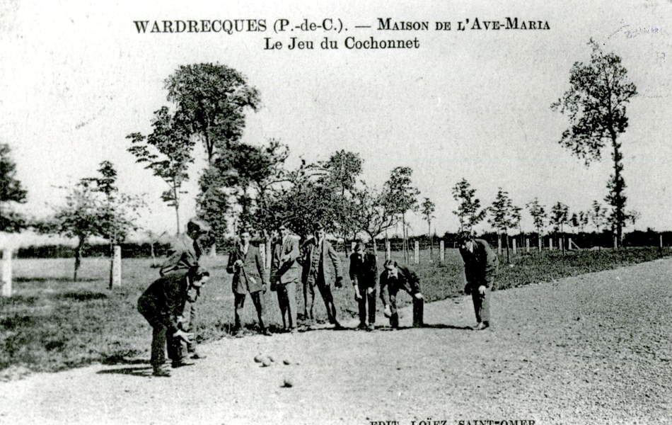 Carte postale noir et blanc montrant 8 hommes en train de jouer à la pétanque dans une allée de graviers. À l'arrière plan, une prairie bordée d'arbres.